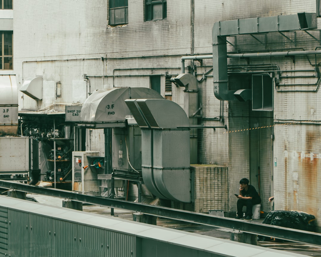 A young man sits on a bucket with his friend in the rooftop mechanical area of a Hong Kong shopping mall, surrounded by HVAC systems, exhaust vents, and stained walls, holding a cigarette during what appears to be his smoke break.