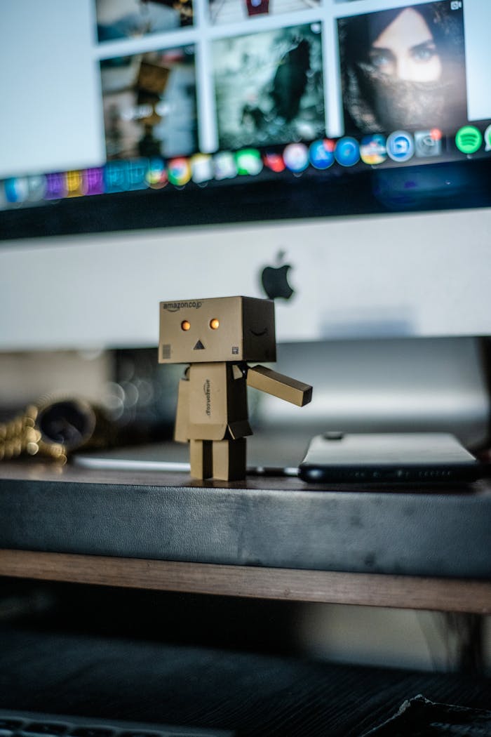 Cardboard robot toy stands on desk with computer screen backdrop, creating a tech and play ambiance.